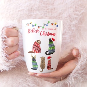 May include: White ceramic mug with a Christmas design featuring cats wearing sweaters. The mug reads "Believe in the magic of Christmas" with a string of colorful lights above the text.