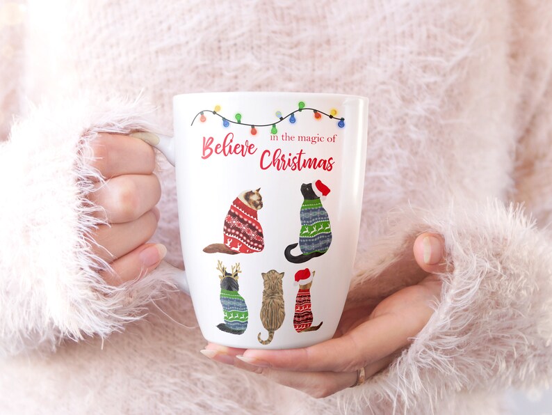 May include: White ceramic mug with a Christmas design featuring cats wearing sweaters. The mug reads "Believe in the magic of Christmas" with a string of colorful lights above the text.
