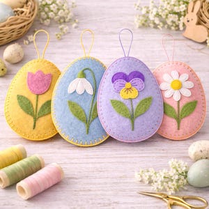May include: Four felt Easter egg ornaments in yellow, blue, purple, and pink, each with a different flower design: a tulip, snowdrop, pansy, and daisy. Each egg has a matching loop for hanging. Decorative spools of thread and scissors are also visible.