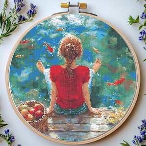 May include: A cross-stitch embroidery of a woman sitting on a wooden dock overlooking a blue lake with red fish swimming. The woman is wearing a red shirt and blue jeans. A basket of fruit sits on the dock beside her.