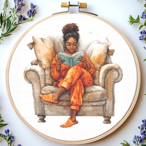 Puede incluir: Una mujer en pijama naranja se sienta en un sillón de color crema y lee un libro azul. Tiene el pelo castaño oscuro y lleva zapatillas.