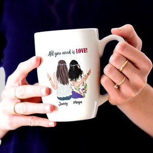 May include: White ceramic mug with a colorful illustration of two women with long hair and floral clothing. The mug has the text "All you need is LOVE!" and the names "Jenny" and "Maya" printed on it.