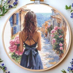 Op de afbeelding: Kruissteekborduurwerk in een houten hoepel toont een vrouw in een blauwe jurk die een geplaveide straat afloopt naar de zee. De scène omvat gebouwen, roze bloemen en blauwe luiken. De borduurwerk is omlijst door een houten hoepel.