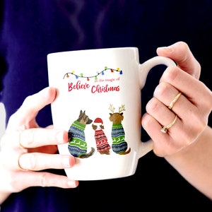 May include: White ceramic mug with a Christmas-themed design featuring three dogs wearing sweaters. The mug has a handle and the text "Believe in the magic of Christmas" is printed on the front.