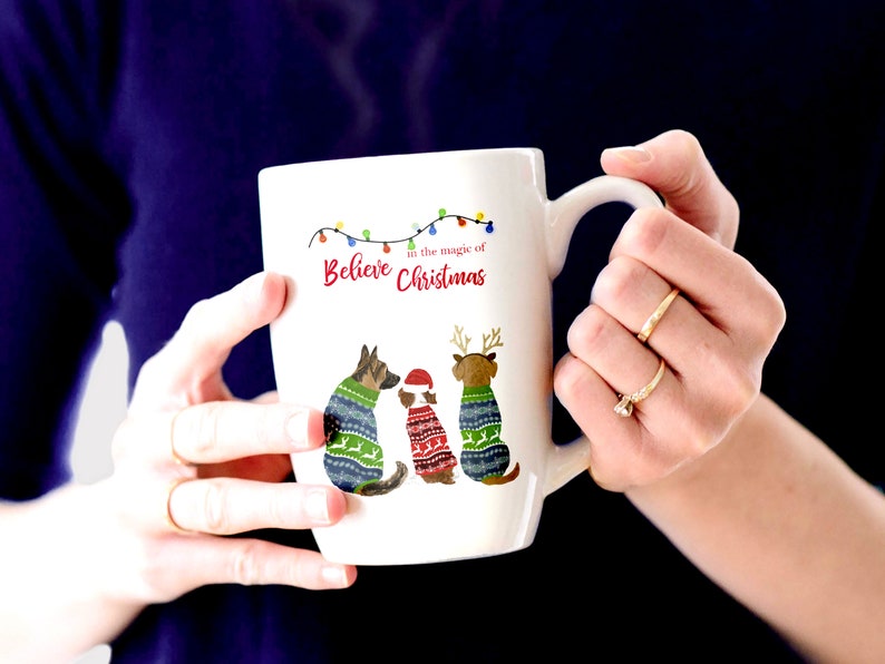 May include: White ceramic mug with a Christmas-themed design featuring three dogs wearing sweaters. The mug has a handle and the text "Believe in the magic of Christmas" is printed on the front.
