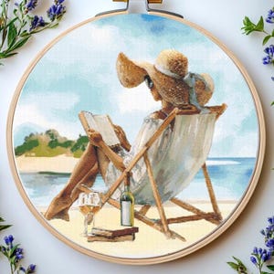 May include: Cross-stitch embroidery in a wooden hoop depicts a person relaxing on a beach, reading a book. The scene includes a beach chair, a bottle of wine, a glass, and a straw hat. The background shows the ocean and sky.