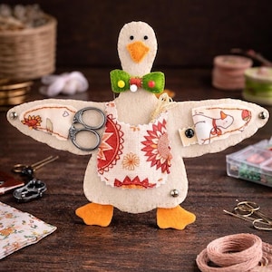 Puede incluir: Un caprichoso organizador de costura con forma de ganso de fieltro. El ganso tiene un cuerpo color crema, patas naranjas y una pajarita verde. Cuenta con un delantal estampado y alas con bolsillos que sostienen tijeras y otros utensilios de costura. El fondo es una superficie de madera oscura.