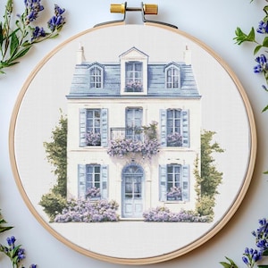 May include: A cross-stitch pattern of a white house with blue shutters and a balcony. The house is surrounded by purple flowers and green bushes.