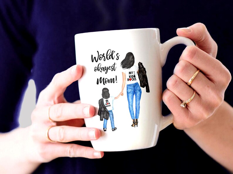 Op de afbeelding: Een witte keramische mok met een zwart-witte illustratie van een moeder en kind. De tekst op de mok luidt "World's okayest Mom!" en "My 1 Kid Rocks".