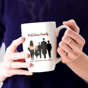 Może przedstawiać: Biały kubek ceramiczny z czerwonym napisem "Finkelman Family" i sylwetką pięcioosobowej rodziny idącej. Pod sylwetkami widnieją imiona Rachel, Chloe, David, Eliana i Ava.