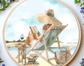 May include: Cross-stitch embroidery in a wooden hoop depicts a person relaxing on a beach, reading a book. The scene includes a beach chair, a glass of wine, a stack of books, and a straw hat. The color palette features blues, tans, and greens.
