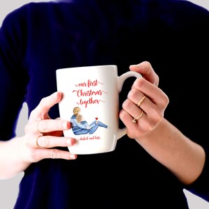 May include: White ceramic mug with a red and blue illustration of a couple in winter clothes. The mug reads "our first christmas together" and "cheers and love".