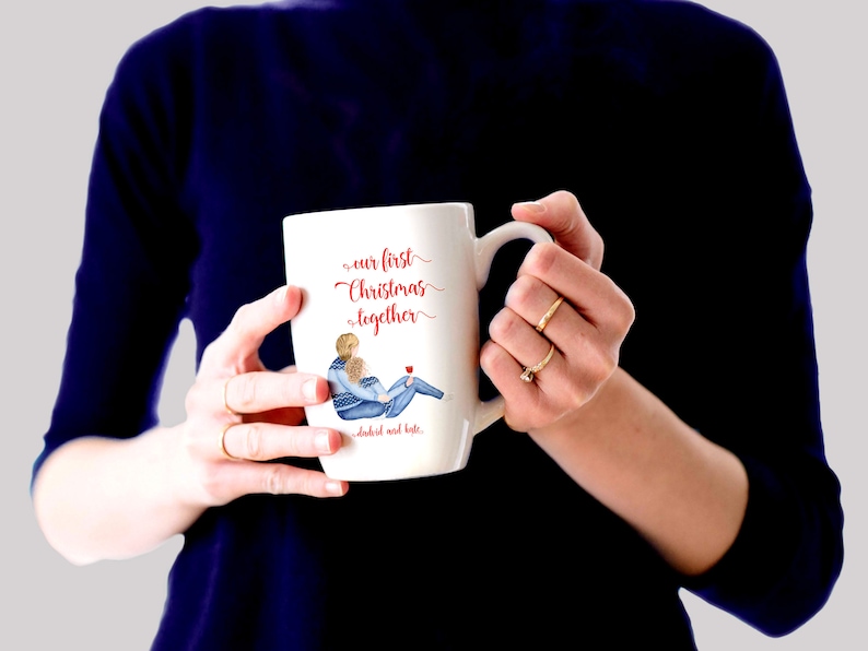 May include: White ceramic mug with a red and blue illustration of a couple in winter clothes. The mug reads "our first christmas together" and "cheers and love".