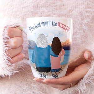 May include: White ceramic mug with a colorful illustration of two women standing back to back on a dock overlooking a lake. The text "The best mom in the WORLD!" is printed on the mug. The names "Doris" and "Dora" are printed below the women.