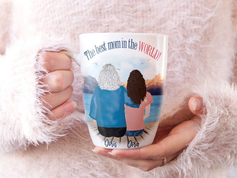 May include: White ceramic mug with a colorful illustration of two women standing back to back on a dock overlooking a lake. The text "The best mom in the WORLD!" is printed on the mug. The names "Doris" and "Dora" are printed below the women.