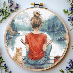Może przedstawiać: Kobieta z długimi brązowymi włosami siedzi nad jeziorem i czyta książkę. Ma na sobie czerwoną koszulę i niebieskie dżinsy. Kot siedzi obok niej. Scena jest namalowana w stylu akwarelowym.