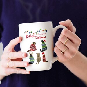May include: White ceramic mug with a Christmas-themed design featuring five cats wearing festive sweaters. The mug has a handle and the text "Believe in the magic of Christmas" is printed on the front.