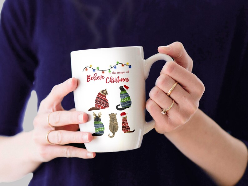 May include: White ceramic mug with a Christmas-themed design featuring five cats wearing festive sweaters. The mug has a handle and the text "Believe in the magic of Christmas" is printed on the front.
