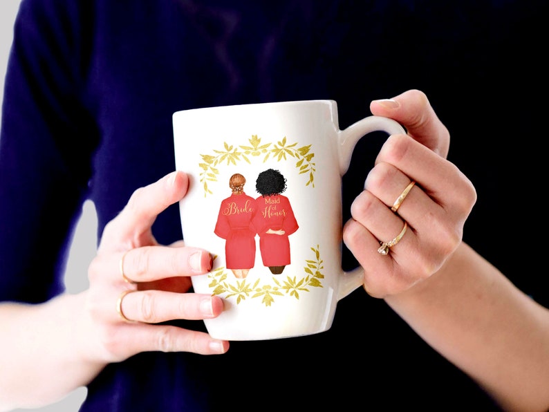 Pode incluir: Caneca de cer&acirc;mica branca com um design floral dourado. Duas mulheres em roup&otilde;es vermelhos s&atilde;o retratadas na caneca, uma com a etiqueta "Bride" e a outra com "Maid of Honor".
