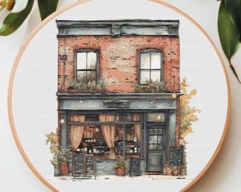 May include: A cross-stitch pattern of a brick building with a storefront. The building has two windows and a door. The storefront has a sign that says "The Bakery". There are plants and flowers in pots outside the building.