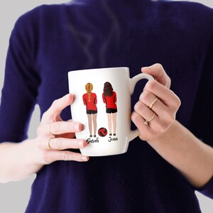 May include: White ceramic mug with a design of two women in red and black volleyball uniforms holding a volleyball. The names "Sarah" and "Jenn" are printed below the women.