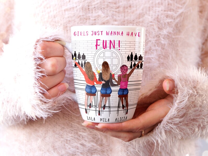 Peut inclure: Mug en c&eacute;ramique blanche avec une illustration color&eacute;e de trois femmes assises &agrave; un comptoir de bar. Le mug porte l'inscription "Girls just wanna have FUN!" en haut et les noms "Lola, Mila, Alicia" en bas.