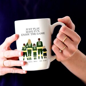 May include: White ceramic coffee mug with a green and black illustration of a family of five wearing hockey jerseys. The text "JUST PLAY. HAVE FUN. ENJOY THE GAME" is printed on the mug. The names "LUKA, MAYA, MILA, JON, JENN" are printed below the illustration.