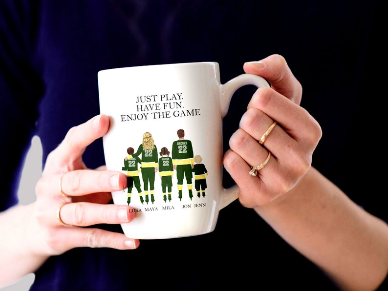 May include: White ceramic coffee mug with a green and black illustration of a family of five wearing hockey jerseys. The text "JUST PLAY. HAVE FUN. ENJOY THE GAME" is printed on the mug. The names "LUKA, MAYA, MILA, JON, JENN" are printed below the illustration.