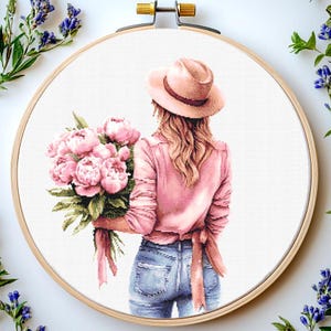 Op de afbeelding: Kruissteekborduurwerk van een vrouw in een roze shirt en hoed, met een boeket roze pioenrozen. De vrouw draagt een blauwe spijkerbroek. De borduursel zit in een houten hoepel, met lavendeltakken op de achtergrond.