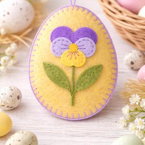 May include: A yellow felt Easter egg ornament with a purple and yellow flower applique. The egg is decorated with a stitched border and a purple hanging loop. Decorative eggs and a basket are in the background.