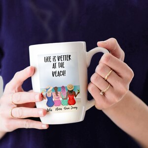 May include: White ceramic mug with a colourful illustration of four women on the beach. The text "Life is better at the beach!" is printed on the mug. The names "Lola, Nina, Sam, Jenny" are printed below the illustration.