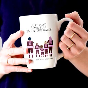 Op de afbeelding: Een witte keramische mok met een roze en bruine illustratie van een gezin van vijf personen die hockeytruien dragen. De tekst "JUST PLAY. HAVE FUN. ENJOY THE GAME" staat op de mok gedrukt. De namen "LUKA, JON, MAYA, MILA, JENN" staan onder de illustratie gedrukt.