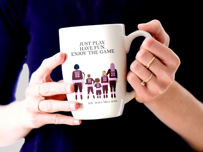 Op de afbeelding: Een witte keramische mok met een roze en bruine illustratie van een gezin van vijf personen die hockeytruien dragen. De tekst "JUST PLAY. HAVE FUN. ENJOY THE GAME" staat op de mok gedrukt. De namen "LUKA, JON, MAYA, MILA, JENN" staan onder de illustratie gedrukt.