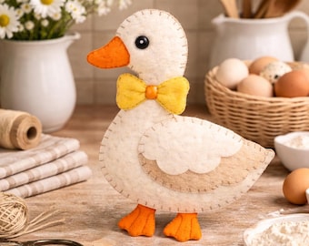 Puede incluir: Un pato de fieltro hecho a mano con pajarita amarilla y pico y patas naranjas. El pato es de color crema con detalles de alas beige y ojos negros. Es un artículo decorativo.