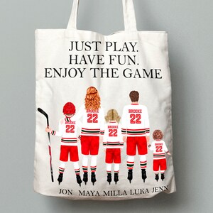 May include: A white canvas tote bag with the text "JUST PLAY. HAVE FUN. ENJOY THE GAME" printed on the front. The bag features a family of five wearing hockey jerseys with the number 22 and the name "BROOKE" printed on the back. The names "JON, MAYA, MILLA, LUKA, JENN" are printed at the bottom of the bag.