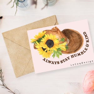 May include: A greeting card with a watercolor illustration of a brown saddle and sunflowers. The card has the text "ALWAYS STAY HUMBLE & KIND" and a brown envelope. The card is on a white surface with glasses and flowers.