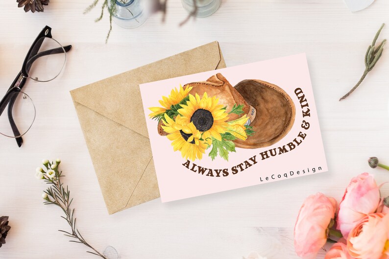 May include: A greeting card with a watercolor illustration of a brown saddle and sunflowers. The card has the text "ALWAYS STAY HUMBLE & KIND" and a brown envelope. The card is on a white surface with glasses and flowers.