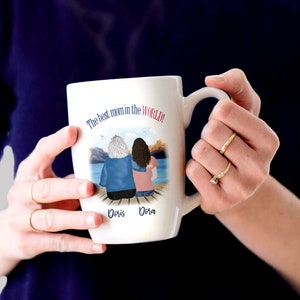 May include: White ceramic mug with a colorful illustration of two women sitting on a dock. The text "The best mom in the world!" is printed on the mug. The names "Doris" and "Dora" are printed below the illustration.