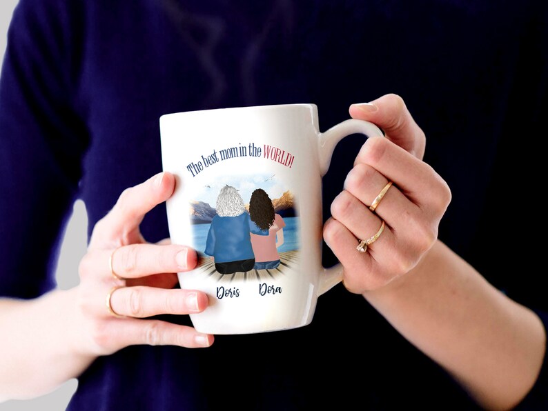May include: White ceramic mug with a colorful illustration of two women sitting on a dock. The text "The best mom in the world!" is printed on the mug. The names "Doris" and "Dora" are printed below the illustration.