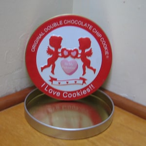 May include: A round, red and white cookie tin with the text "ORIGINAL DOUBLE CHOCOLATE CHIP COOKIE" and "I Love Cookies!!". The lid features two red cherubs holding a heart. The tin is open, revealing a silver interior.