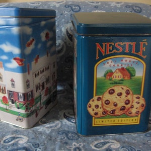 May include: Two rectangular metal cookie tins. One tin features a blue sky with clouds and falling leaves, and a detailed illustration of a house. The other tin is blue with the Nestle Toll House Cookies logo and images of chocolate chip cookies.