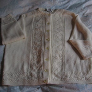 May include: Cream-colored cardigan sweater with a button-up front. The sweater features a textured diamond pattern down the center and on the bottom hem. The sleeves have a decorative cuff. The buttons are a pale yellow color.