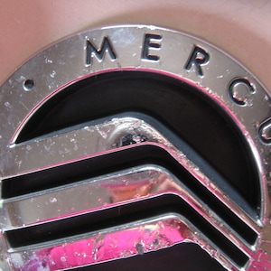 May include: Close-up of a chrome Mercury emblem with the brand name in capital letters. The emblem has a circular shape with a black background and three horizontal lines. The emblem is set against a light pink background.