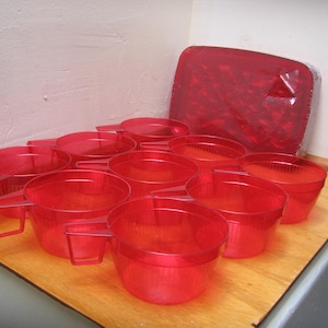 May include: A set of ten translucent red plastic cups with handles, arranged on a wooden surface. A matching red plastic tray is also visible. The cups and tray appear to be part of a kitchen or camping set.