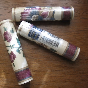 May include: Three rolls of floral wallpaper. The wallpaper features a cream background with purple, blue, and green floral patterns. The bottom border has a dark purple design. The rolls are wrapped in clear plastic.