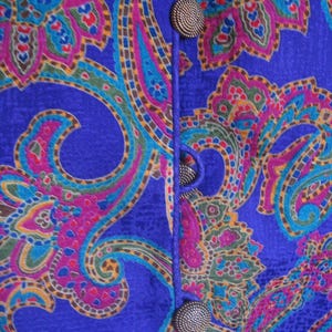 May include: Close-up of a blue fabric with a paisley pattern in shades of pink, teal, green, and yellow. The fabric is fastened with three textured, round, bronze-colored buttons.