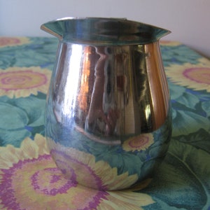 May include: A shiny, silver-colored creamer with a flared spout. The creamer has a rounded body and a slightly wider top. The surface reflects the surrounding environment. The creamer is sitting on a floral patterned tablecloth.