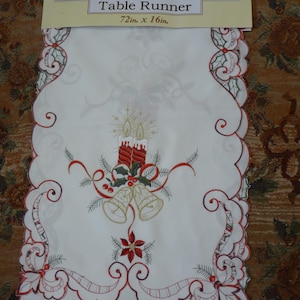 May include: A white decorative table runner with a scalloped edge, measuring 72 inches long and 16 inches wide. It features embroidered Christmas bells, candles, and holly. The text "Decorative Table Runner" is printed on the tag.