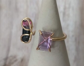 May include: A gold ring with a pink and black gemstone and a purple square gemstone.