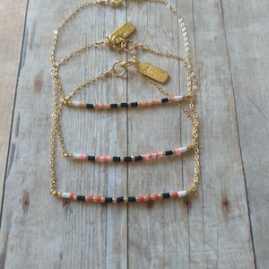 May include: Three gold chain bracelets with small rectangular tags that say "LOVE" on them. Each bracelet has a different colored beaded bar: pink, white, and orange.